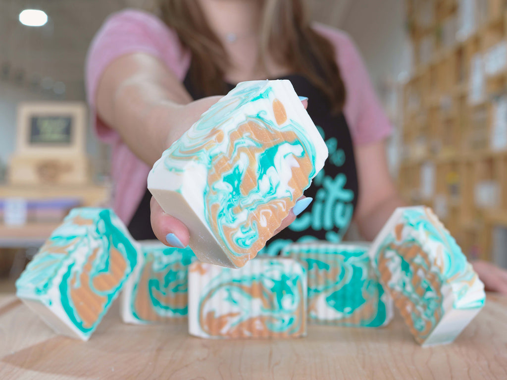 Buff City Soap maker holding up a white, teal and brown soap bar with more of the same soap bars in frame.
