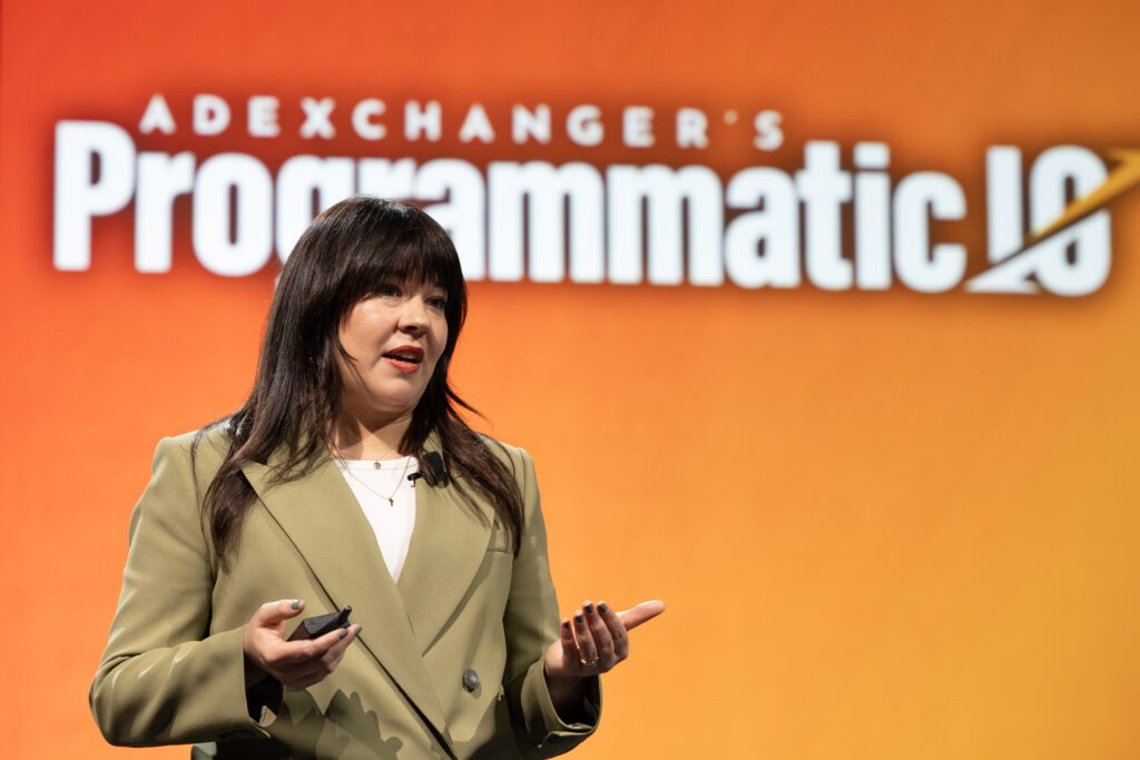 Katie Barrett, Head of Strategic Sales at LG Ads Solutions on stage at AtExchanger's Programmatic IO with a tan blazer infront of an orange background.