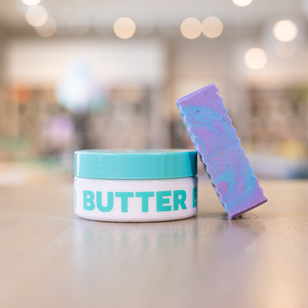 Purple and blue soap bar leaning against clear body butter container with teal lid on counter